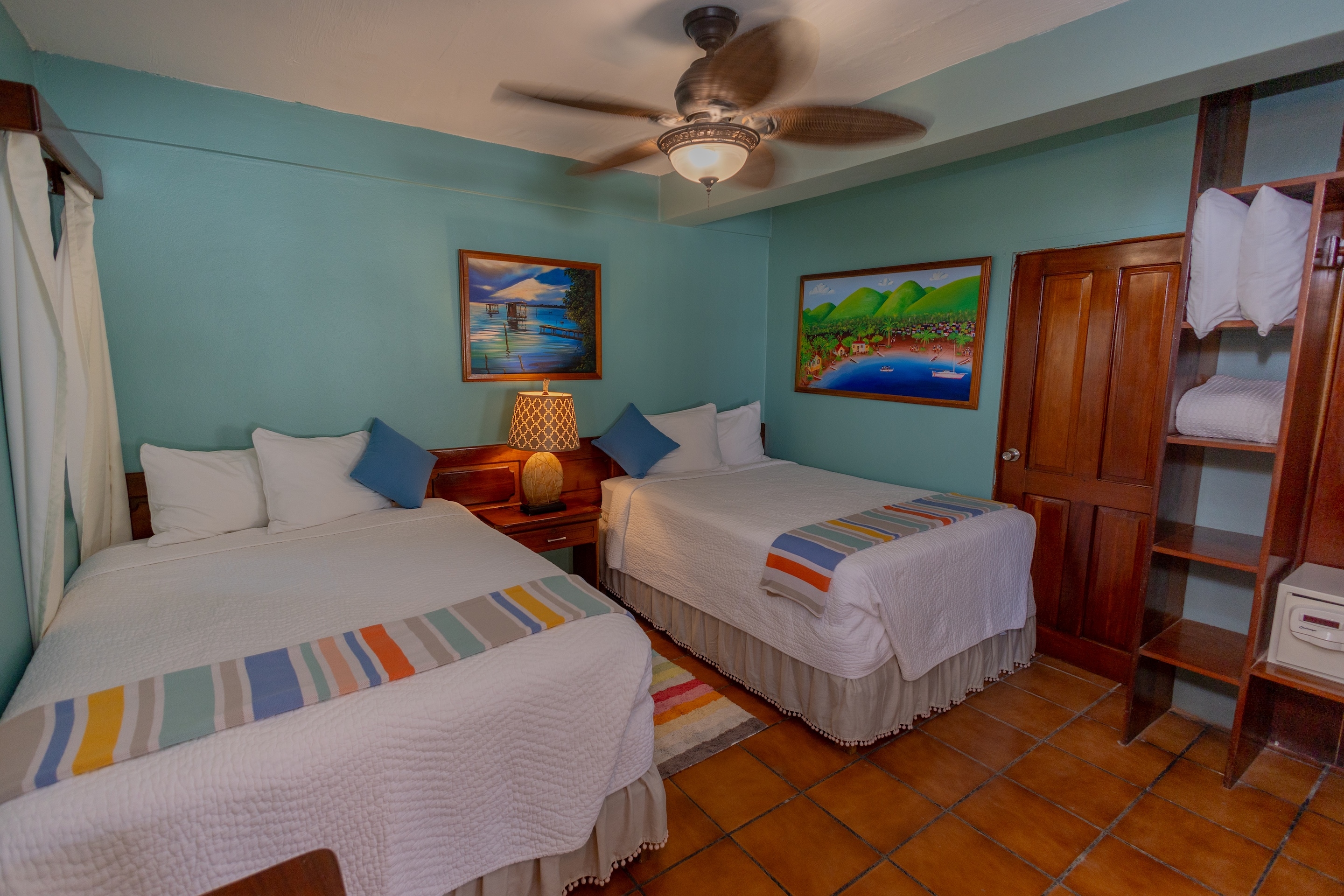 A bedroom with teal walls, tiled floors, two full beds with colorful accents, a ceiling fan, and fra