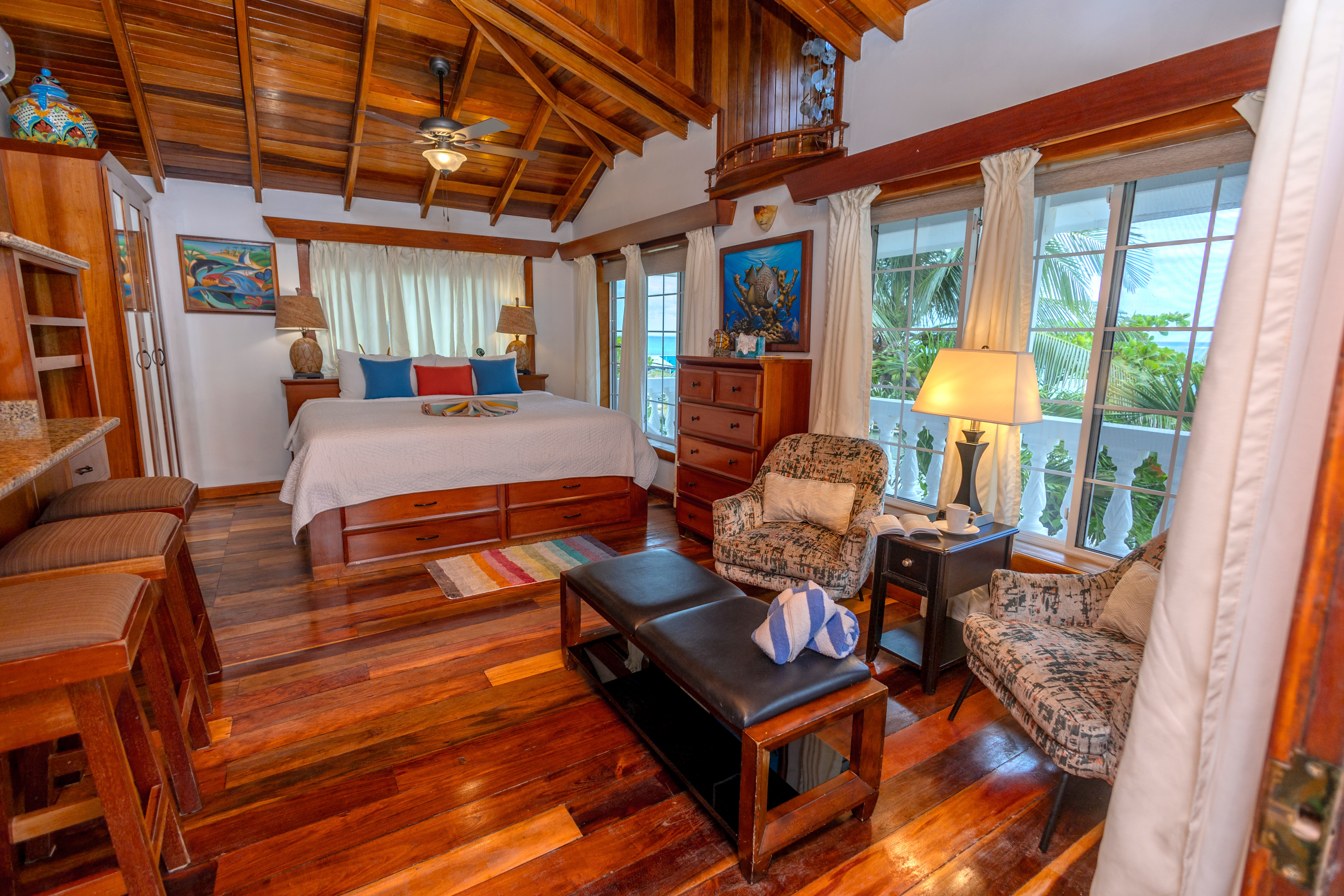 A bedroom with wood paneled walls and ceiling, a bed with colorful pillows, seating area, and balcon