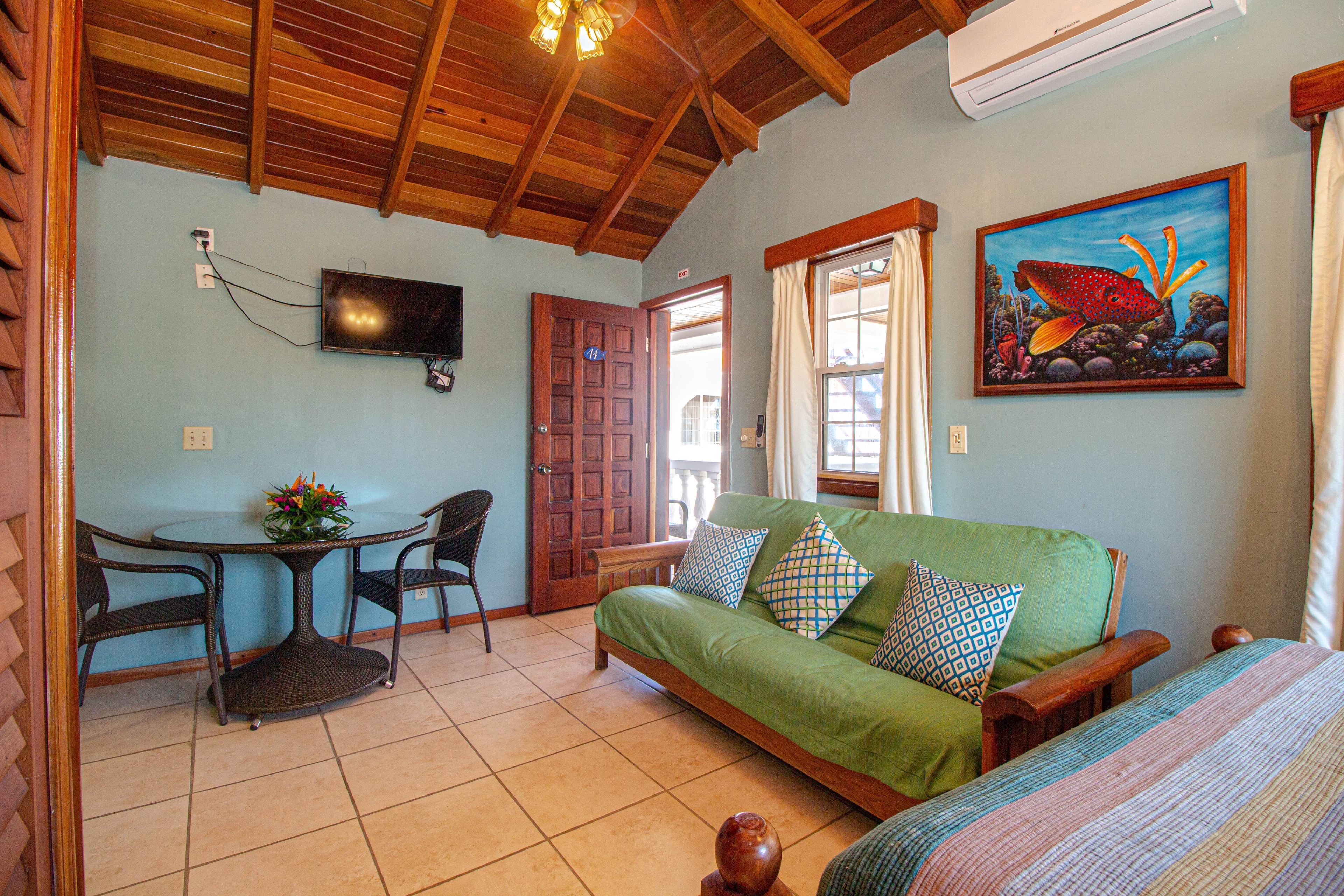 A room with a high wooden ceiling, a green futon, a round table with chairs, a mounted TV, and a pai
