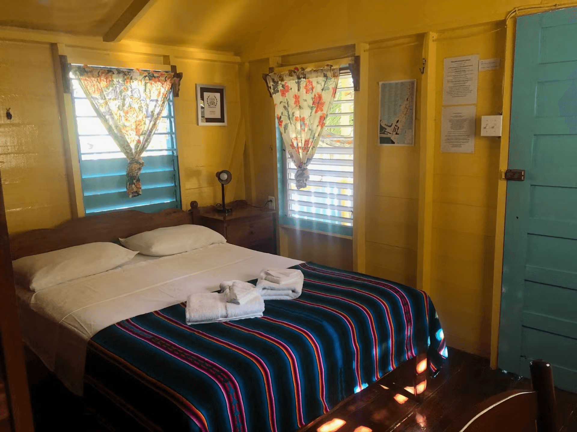 Bedroom with bright yellow walls, double bed with colorful striped blanket and white sheets, sunny w