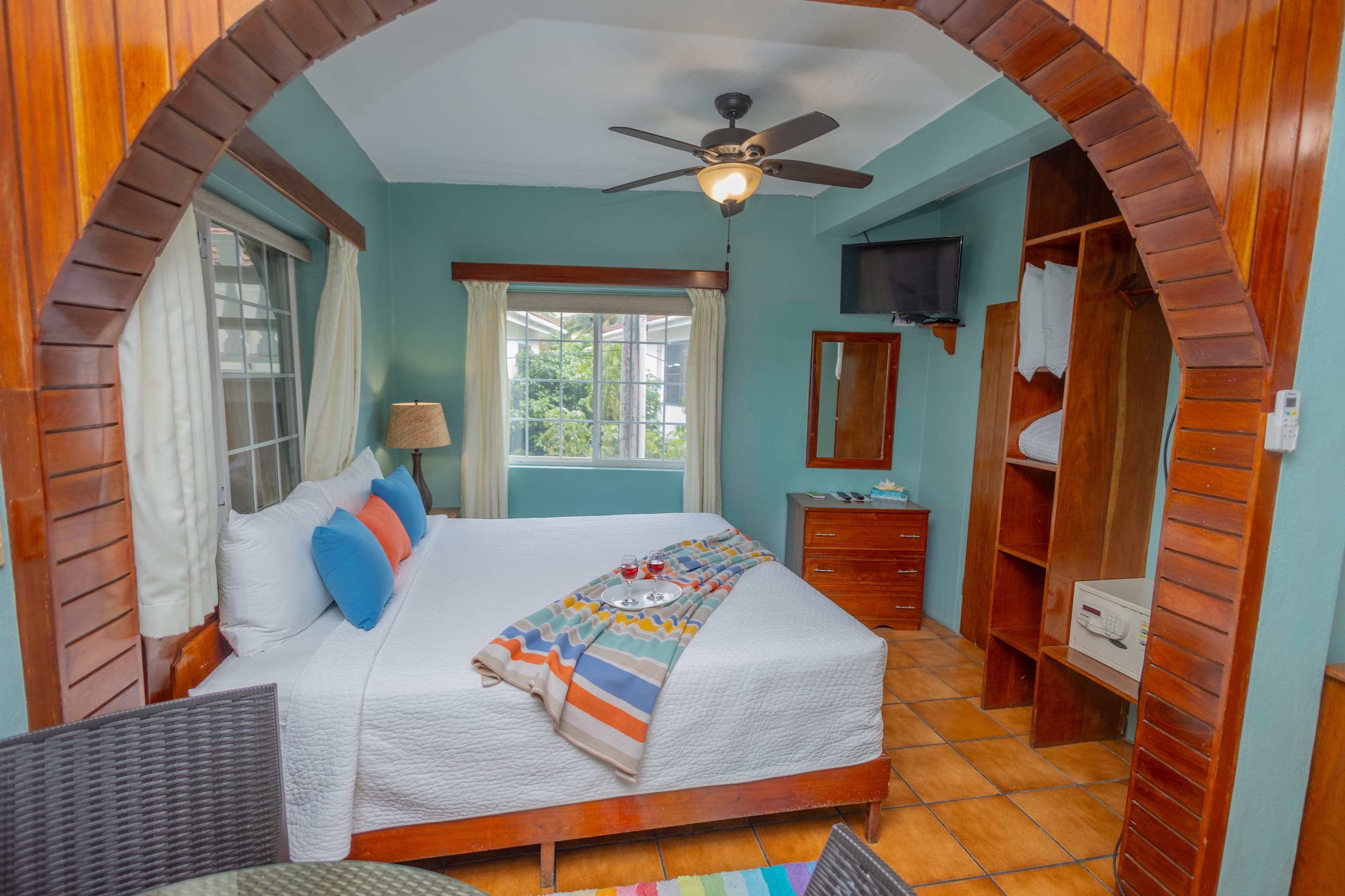 A bedroom with teal walls, tiled floors, a king bed with colorful pillows and runner, a ceiling fan,