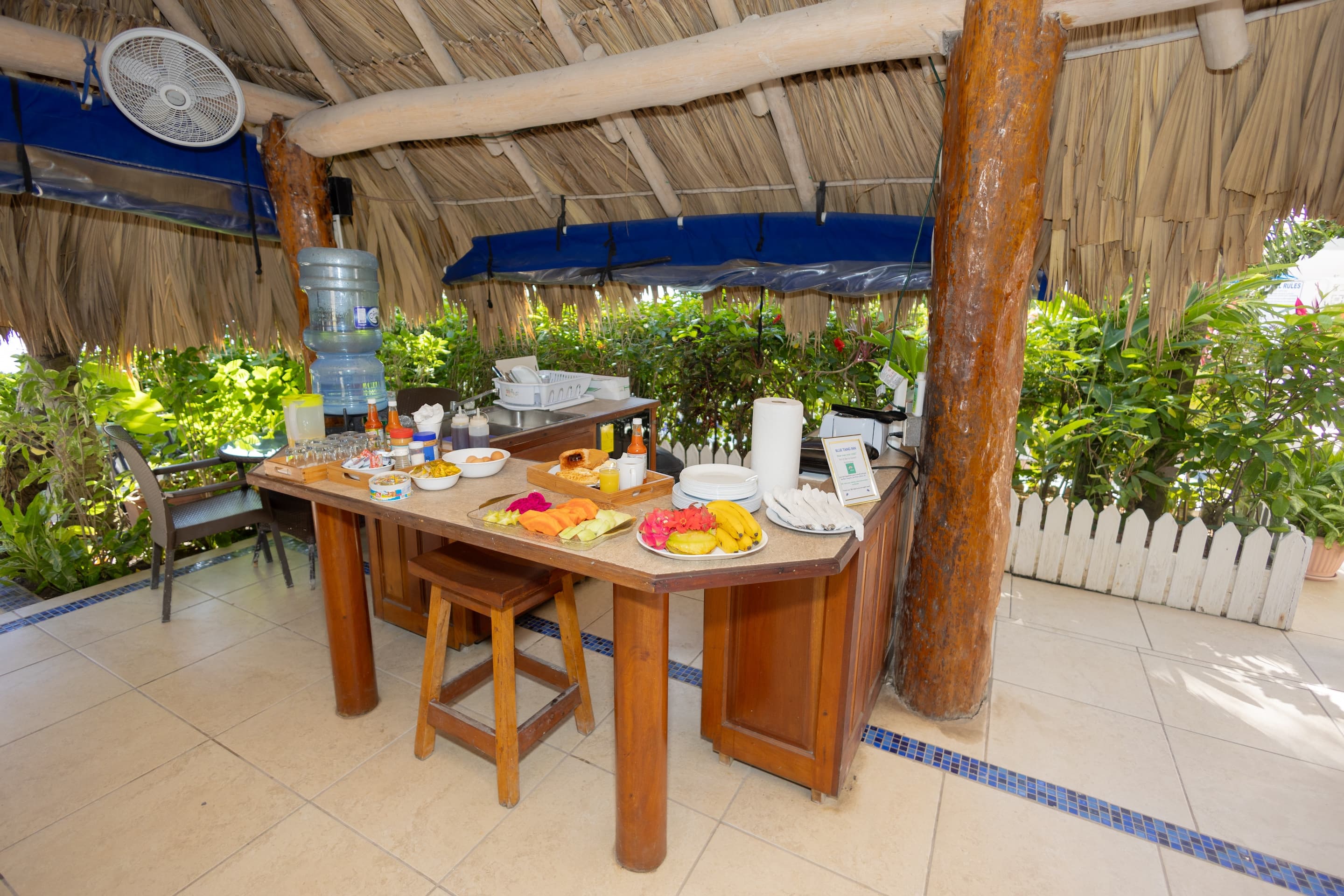 An open-air buffet setup under a thatched roof with food, drinks, and a water cooler, surrounded by