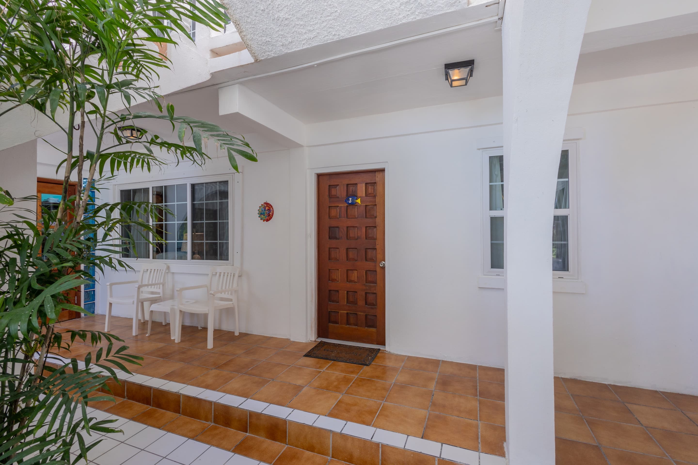 A covered outdoor entrance area with white walls, a wooden door, and tropical plants.
