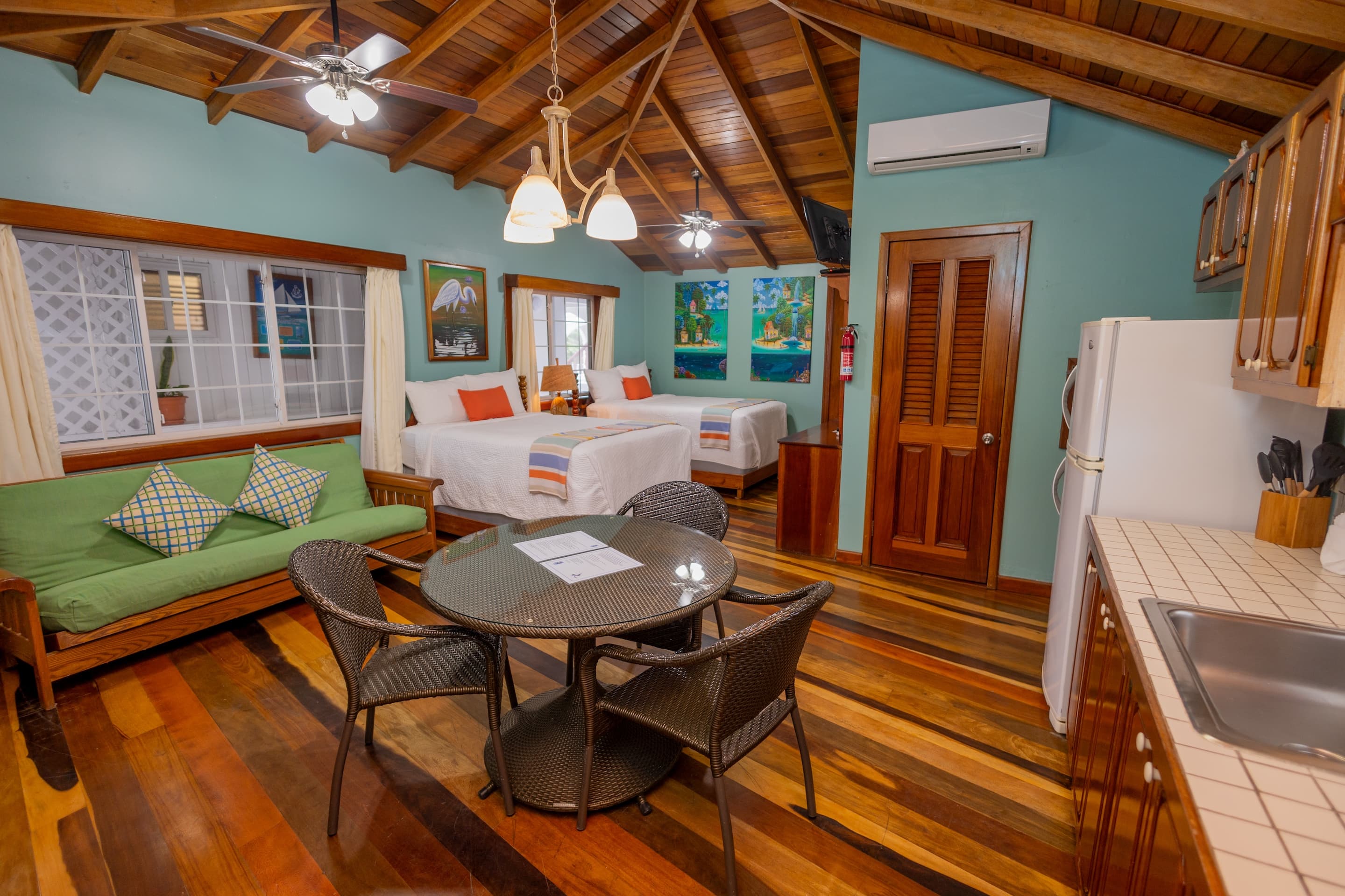 A room with a high wooden ceiling, a green futon, a round table with chairs, two beds, and a kitchen