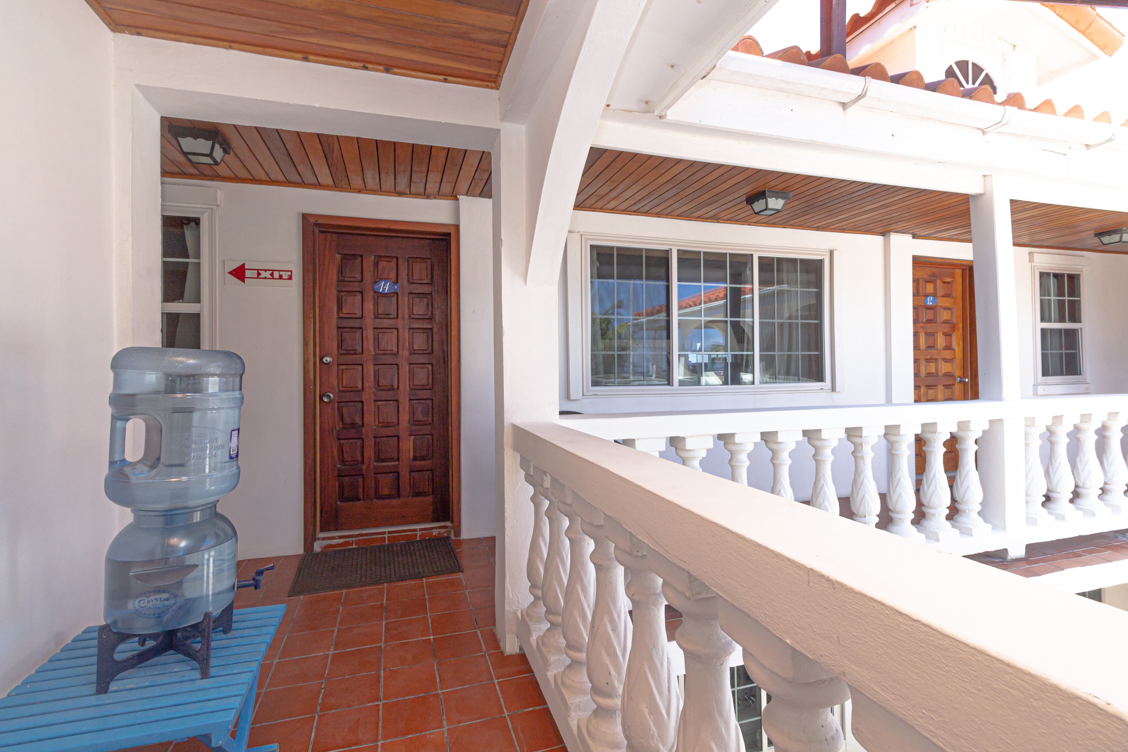 An outdoor balcony with white walls and railings, wooden doors, and a blue water cooler on a table.