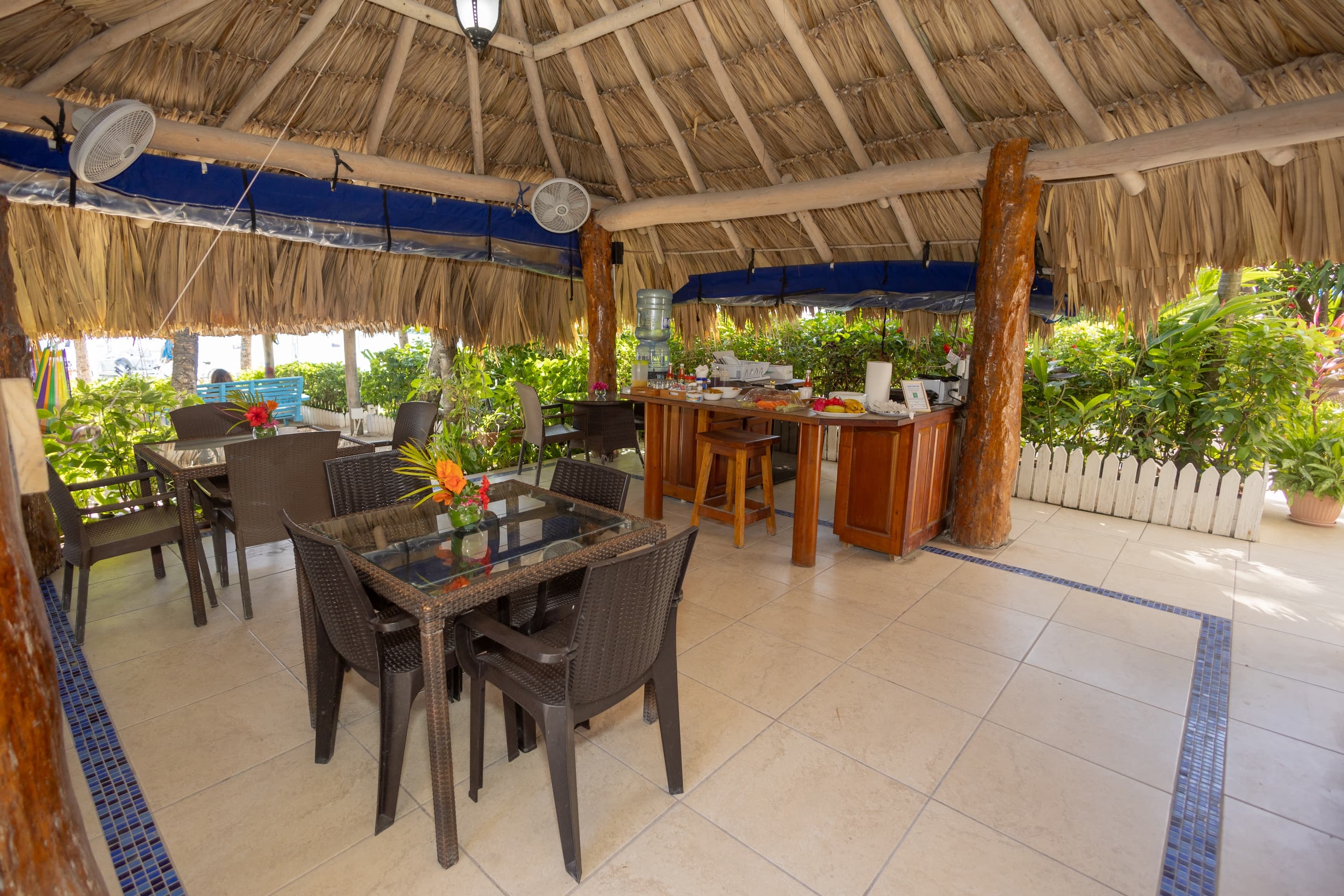 An open-air dining area with a palapa roof, tables and chairs, and a bar, surrounded by greenery.