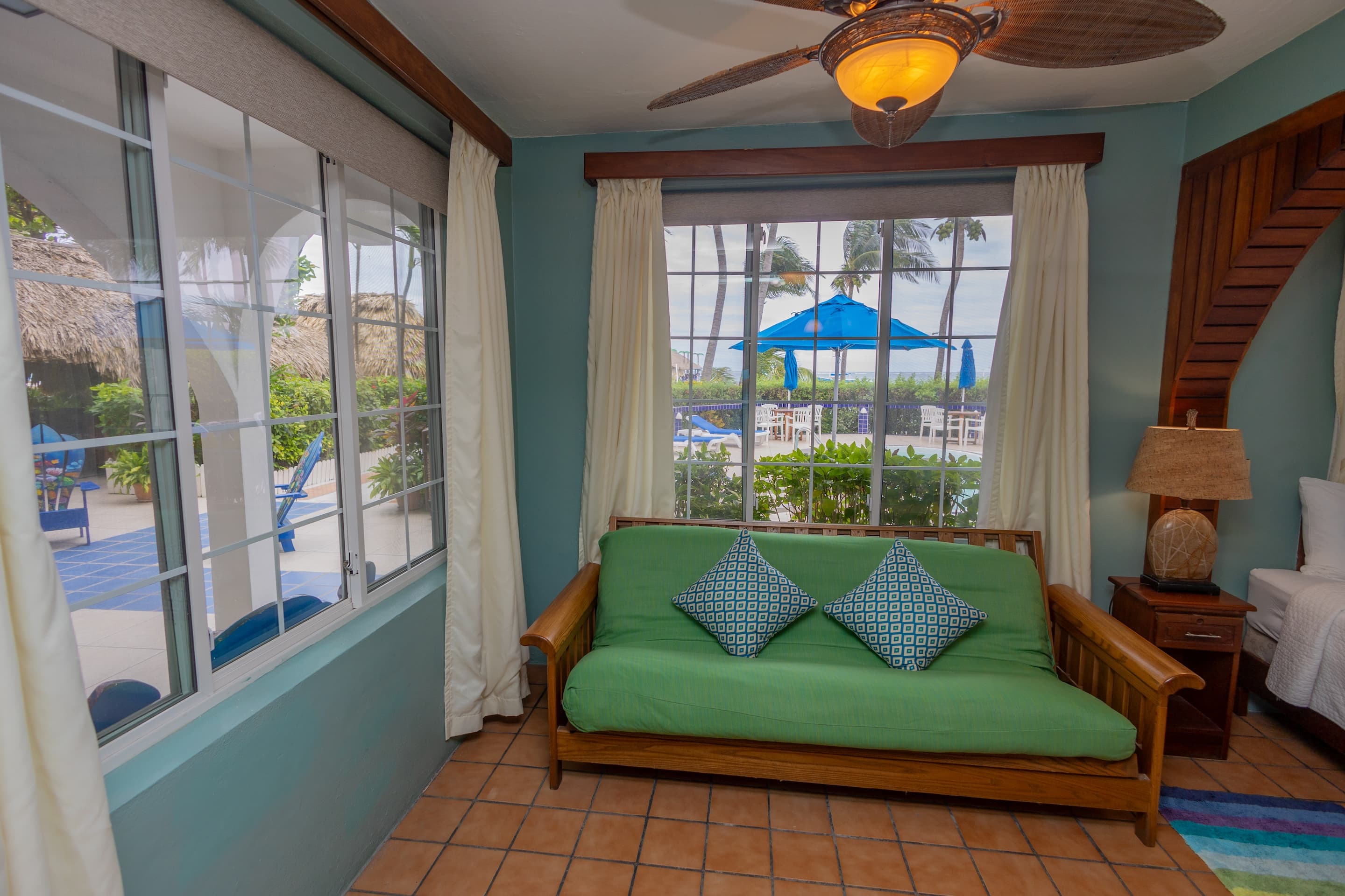 A room with teal walls, tiled floors, a green futon with blue pillows, a ceiling fan, and windows wi