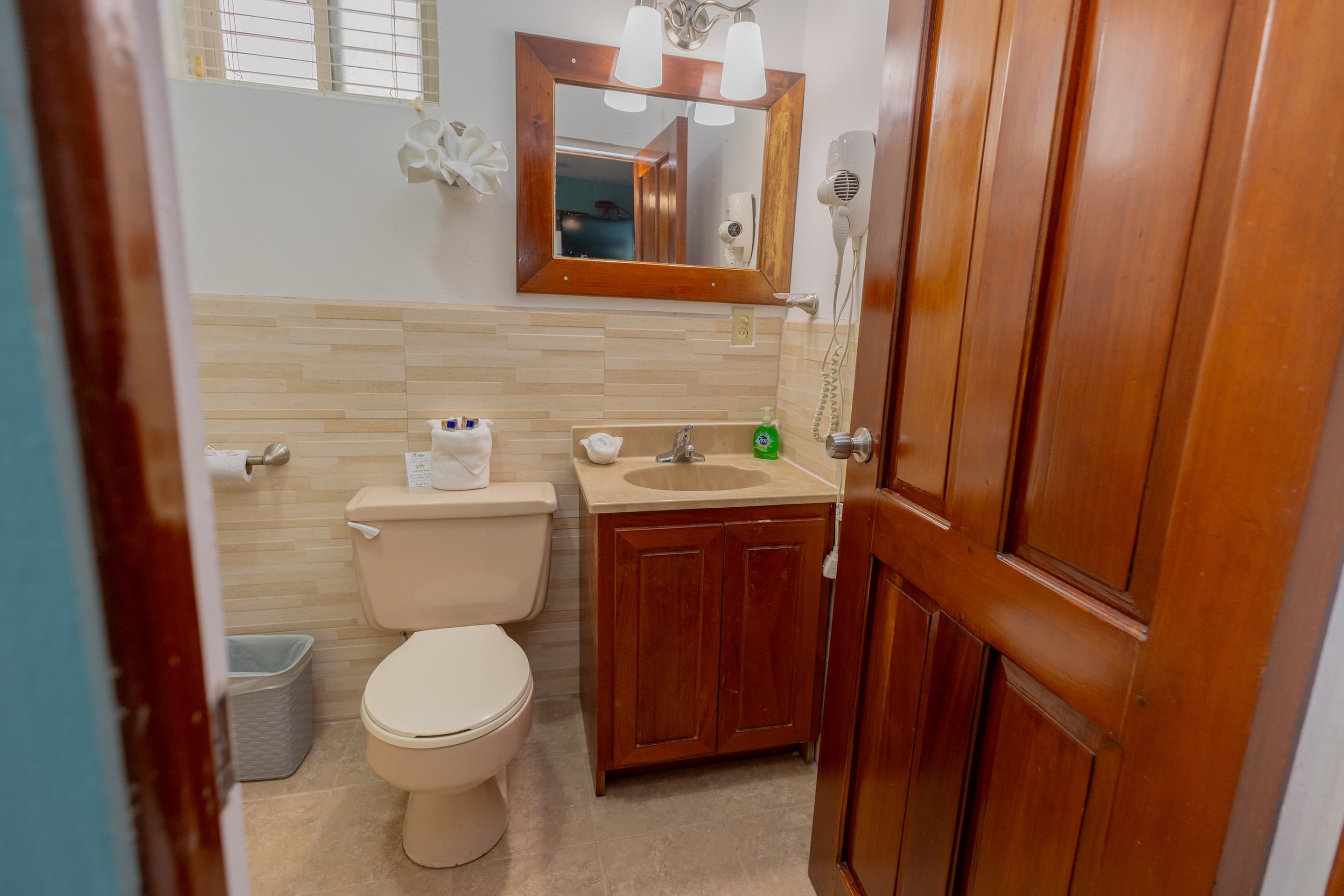 A compact bathroom with light tiled walls, a wooden-framed mirror above a sink with a wooden cabinet