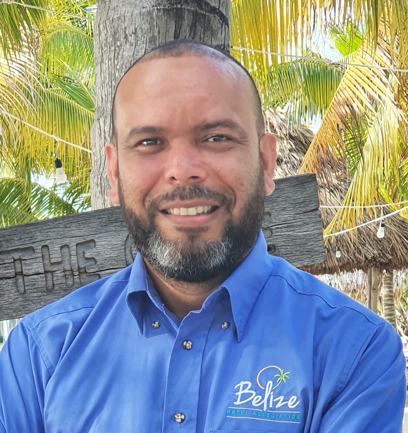 A man with a beard smiles in front of tropical foliage and a wooden sign.