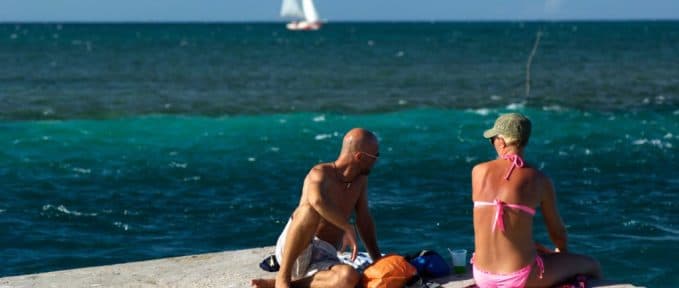 A couple relaxes on a seaside pier while a sailboat floats in the background.