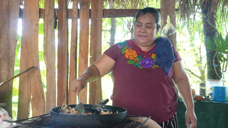 A woman cooks in a large pan over an open fire in a rustic kitchen.