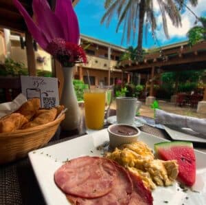 A beautifully arranged breakfast plate with ham, scrambled eggs, a slice of watermelon, and chocolate sauce, accompanied by a juice and coffee, set in a courtyard.