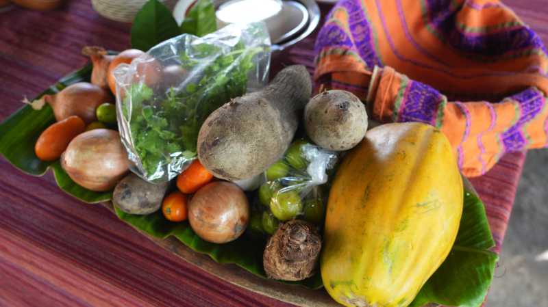 A wooden tray lined with banana leaves holds a variety of fresh vegetables and fruits, including potatoes, tomatoes, and a papaya.