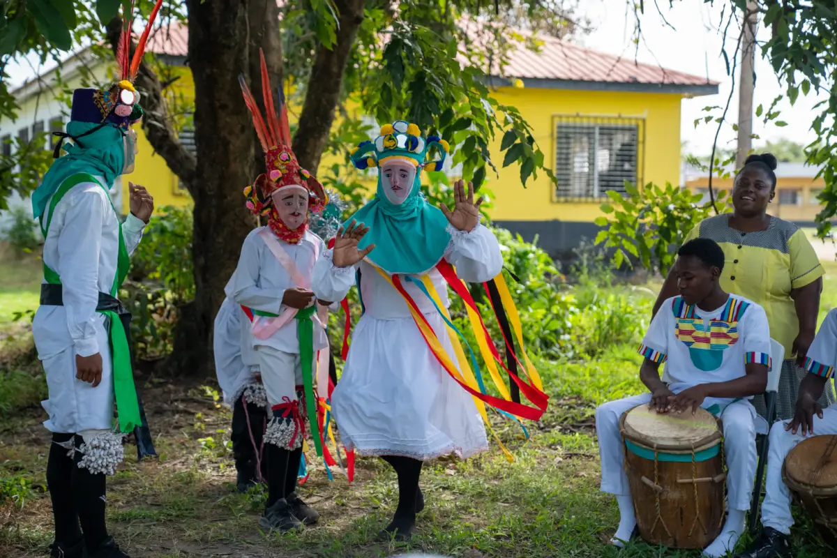 A group of performers in colorful costumes dances outdoors near a yellow building.