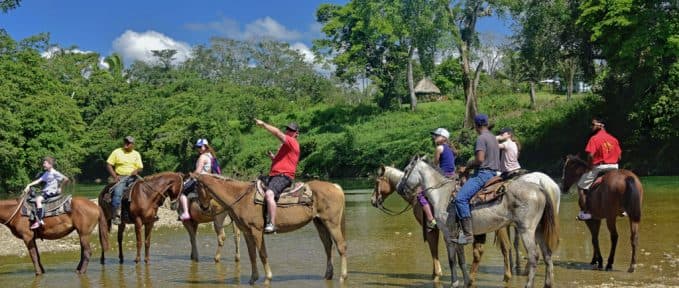 A group of people on horseback stands in a river, surrounded by lush greenery.
