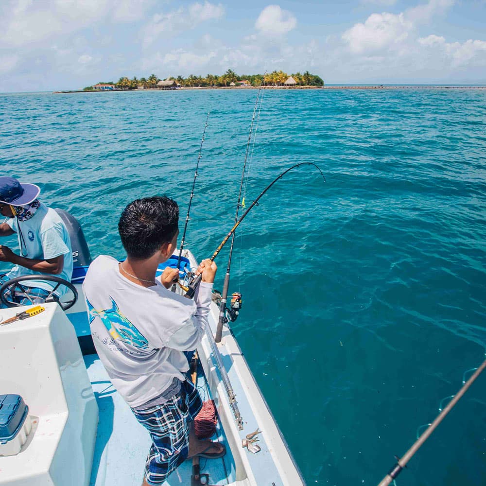 Two fishermen on a boat are fishing in clear blue waters near an island.