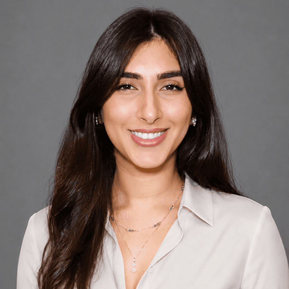A young woman with long dark hair smiles warmly, wearing a beige blouse and delicate jewelry against a gray background.