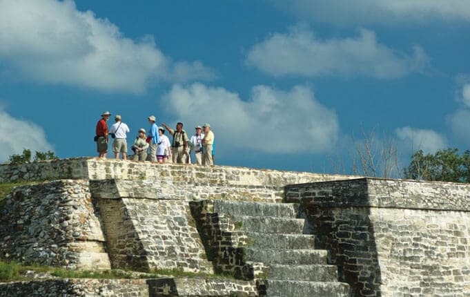 A group of tourists explores an ancient stone structure under a blue sky with clouds.