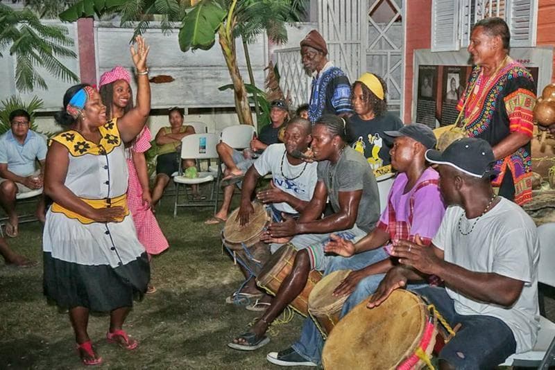 A lively group of musicians and dancers perform together outdoors, surrounded by lush greenery.