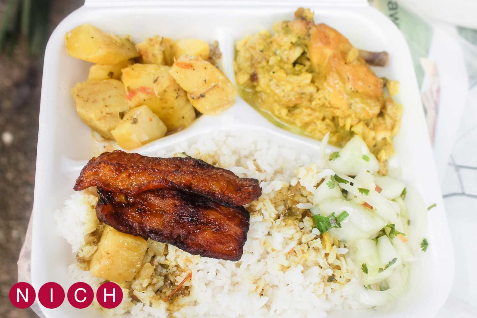 A white takeout container filled with rice, fried plantains, cooked vegetables, and a side of fresh cucumber salad.