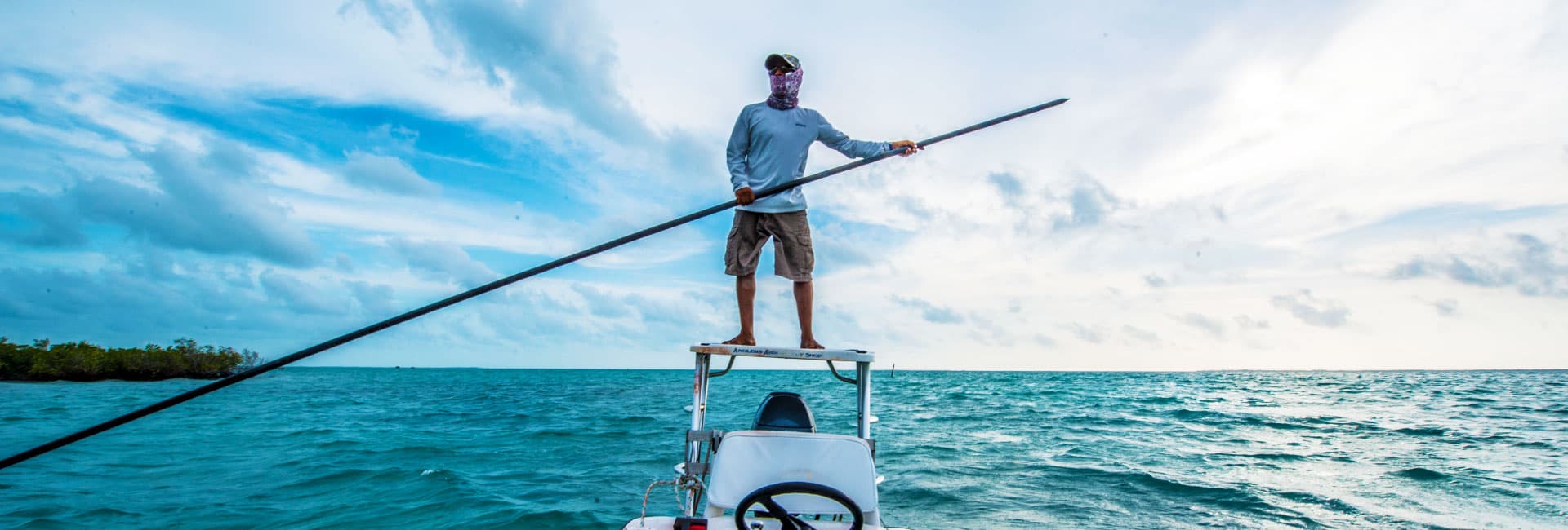 A person stands on a fishing boat, holding a long pole against a backdrop of blue water and cloudy sky.