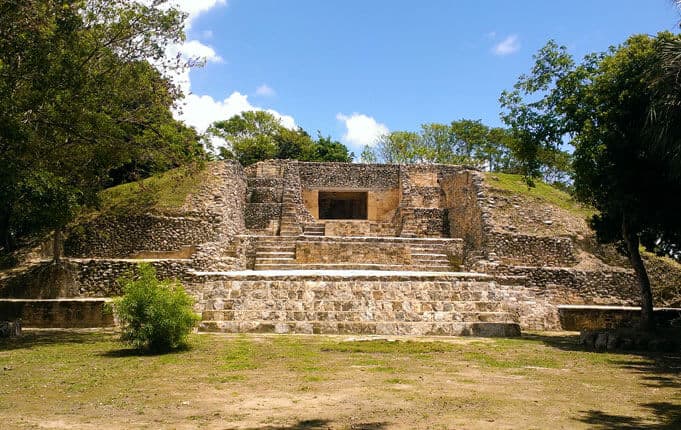 Ancient stone steps lead up to a partially restored pyramid structure surrounded by trees and blue sky.