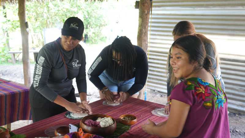 A group of women joyfully prepares traditional food at a table outdoors.
