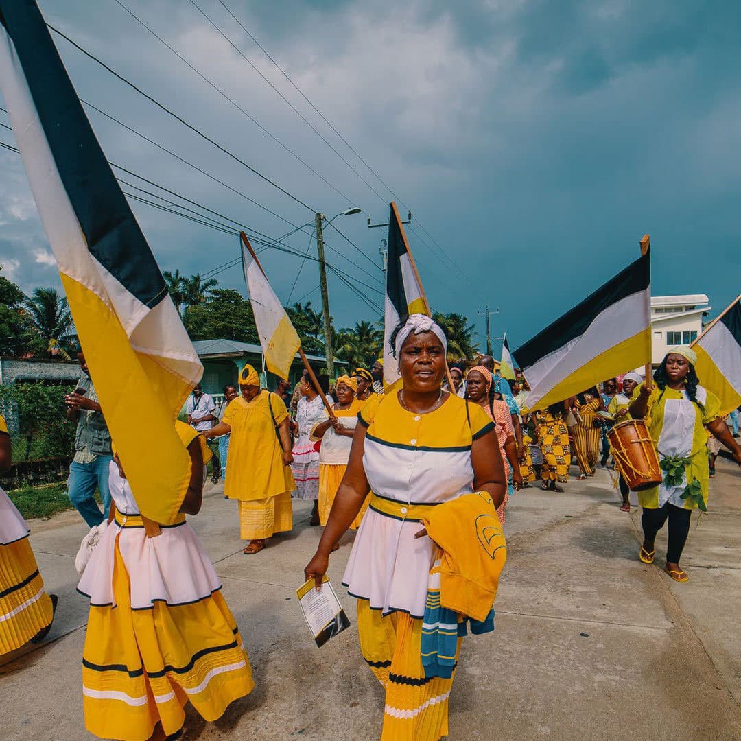 A group of women in vibrant yellow and white attire march while waving flags against a cloudy sky.