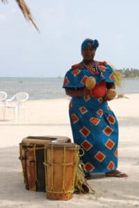 A woman in traditional attire plays maracas near a beach with drums beside her.