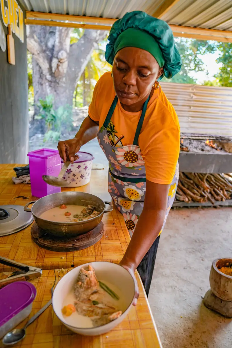 A woman in a colorful apron serves fish soup from a pot in a rustic kitchen.