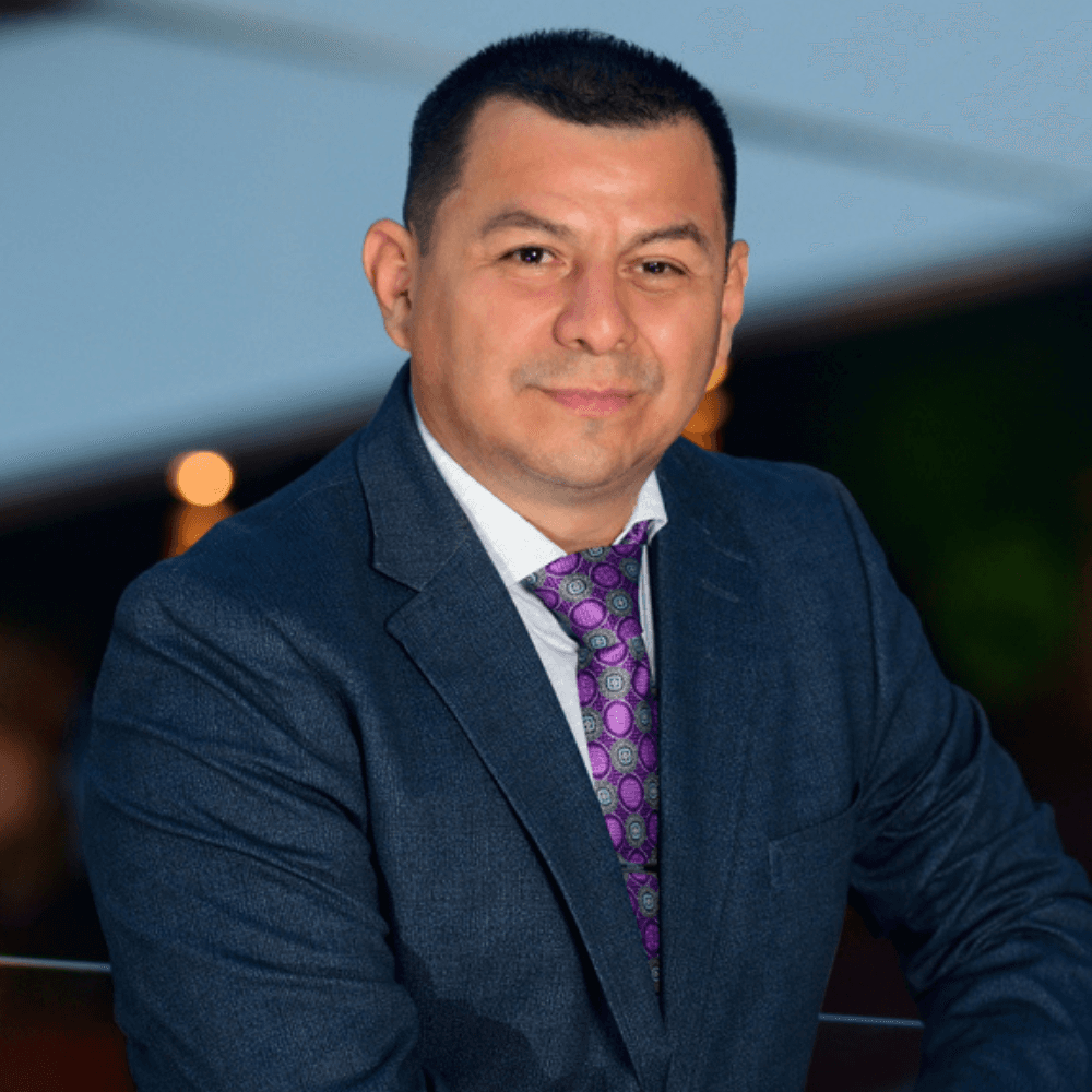 A man in a dark suit and patterned tie poses confidently against a softly blurred background.