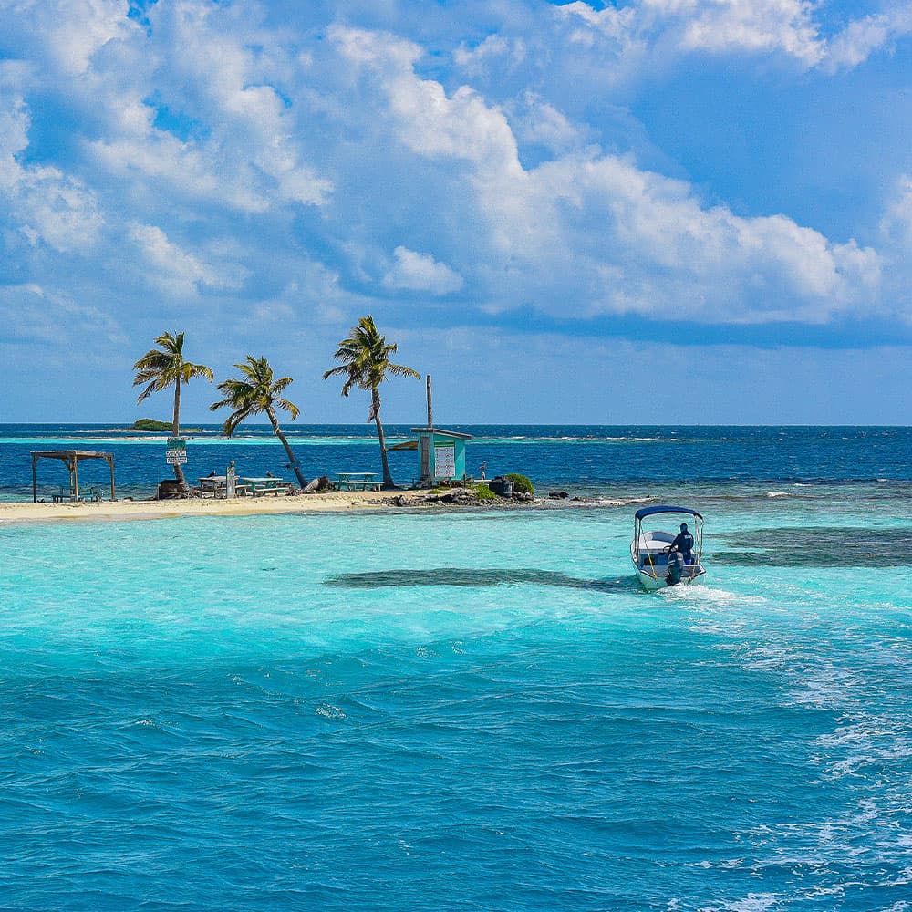 A small boat navigates through turquoise waters near a sandy island with palm trees and a beach hut.