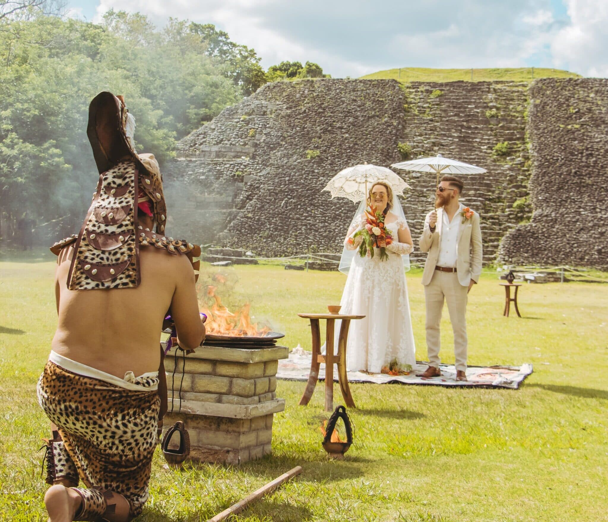 A wedding ceremony taking place outdoors with a couple in formal attire and a person in traditional dress near a fire.