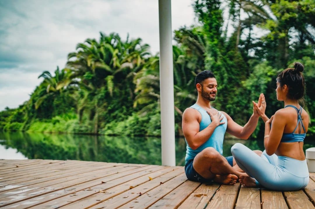 A man and woman practice yoga on a wooden dock beside a serene, lush landscape.