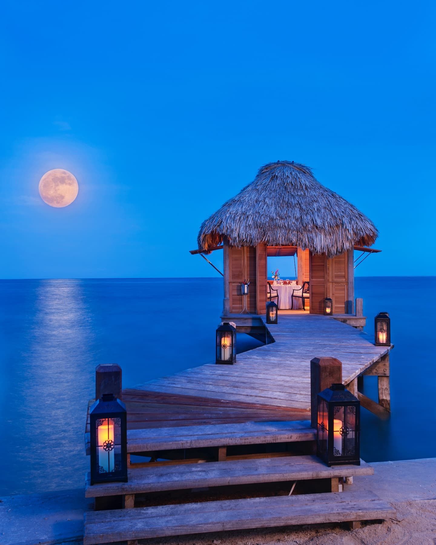 A thatched hut on a pier with lanterns under a full moon over calm water.
