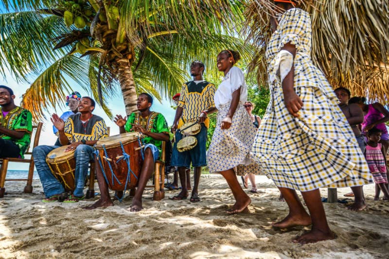 A group of people wearing traditional clothing plays drums and dances on a sandy beach under palm trees.