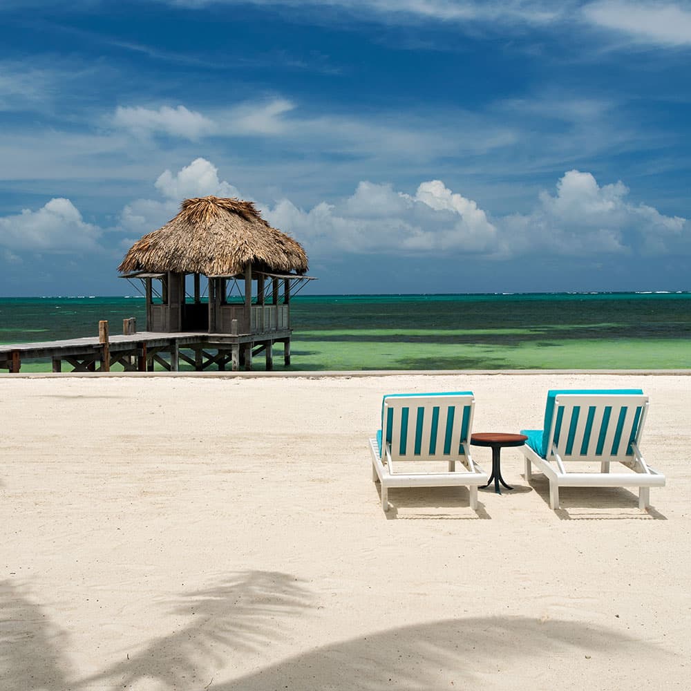 Two striped lounge chairs face a wooden gazebo on a sandy beach by the ocean.